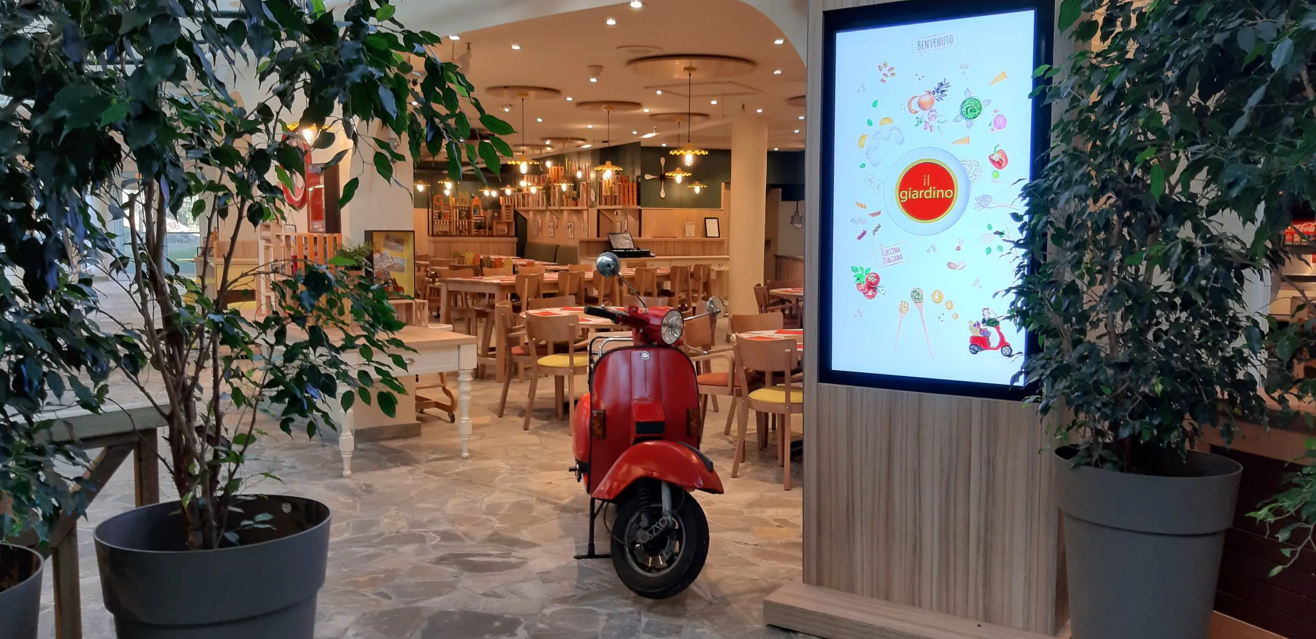 Entrée d’un restaurant avec une décoration végétale et un scooter rouge exposé à l’intérieur.
