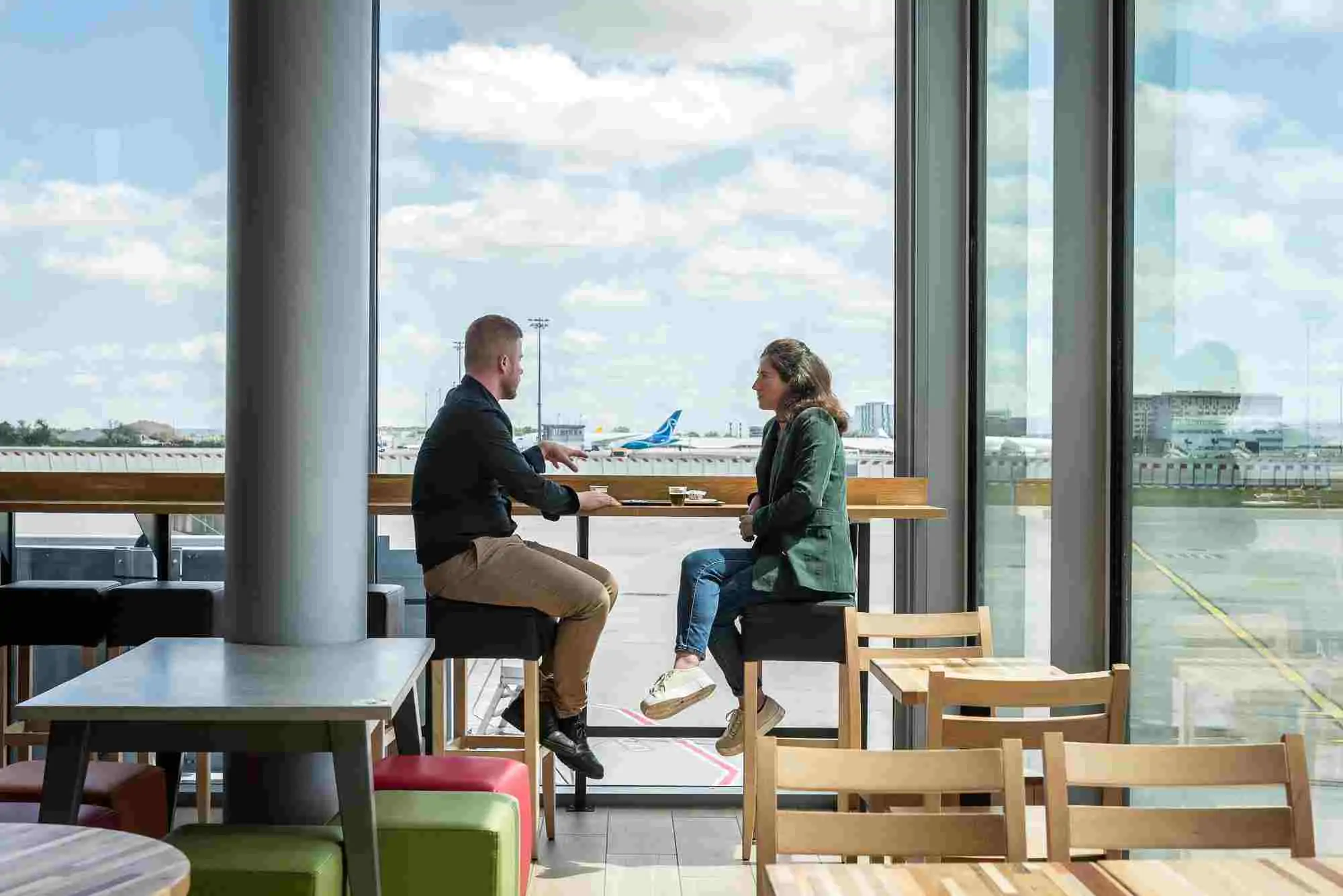 Deux personnes assises à une table haute dans un espace de restauration avec vue sur les pistes d’un aéroport.