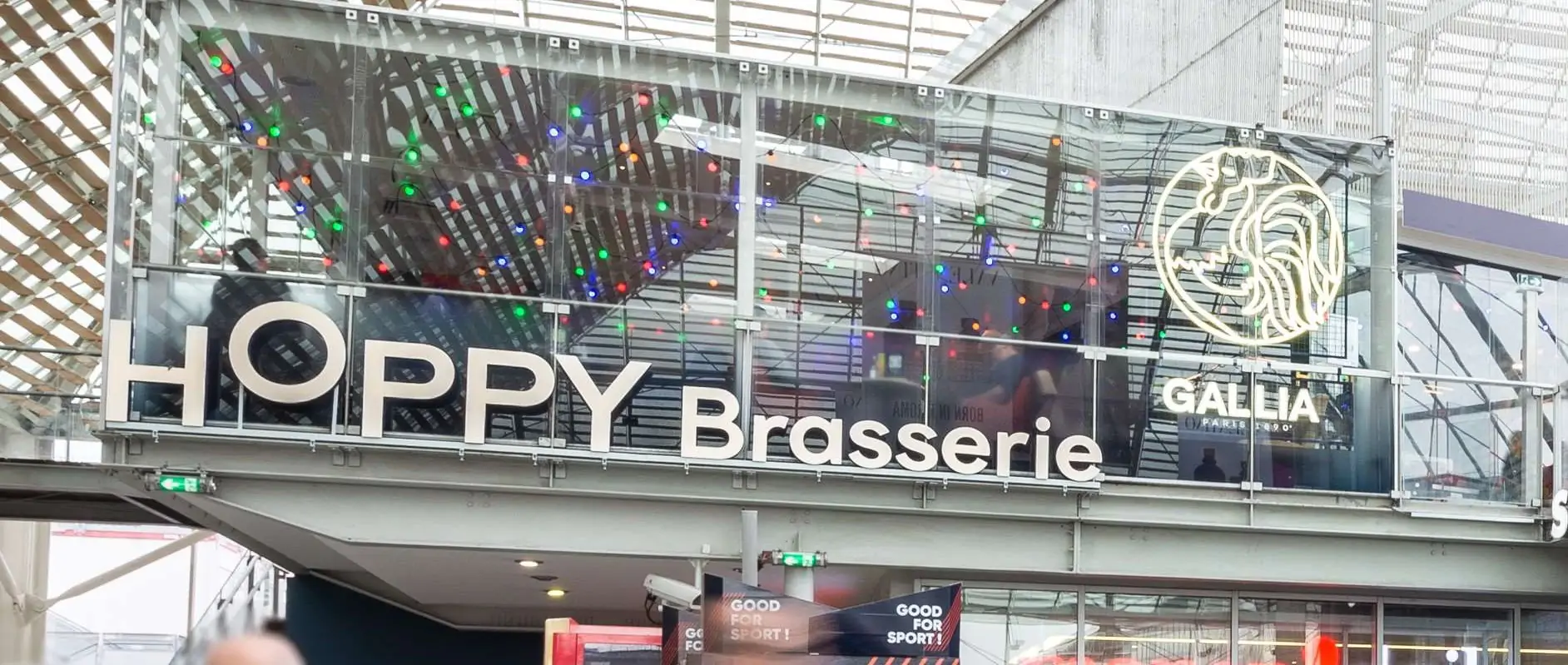 Façade vitrée du restaurant Hoppy Brasserie dans une gare, avec une enseigne lumineuse et des passants en contrebas.